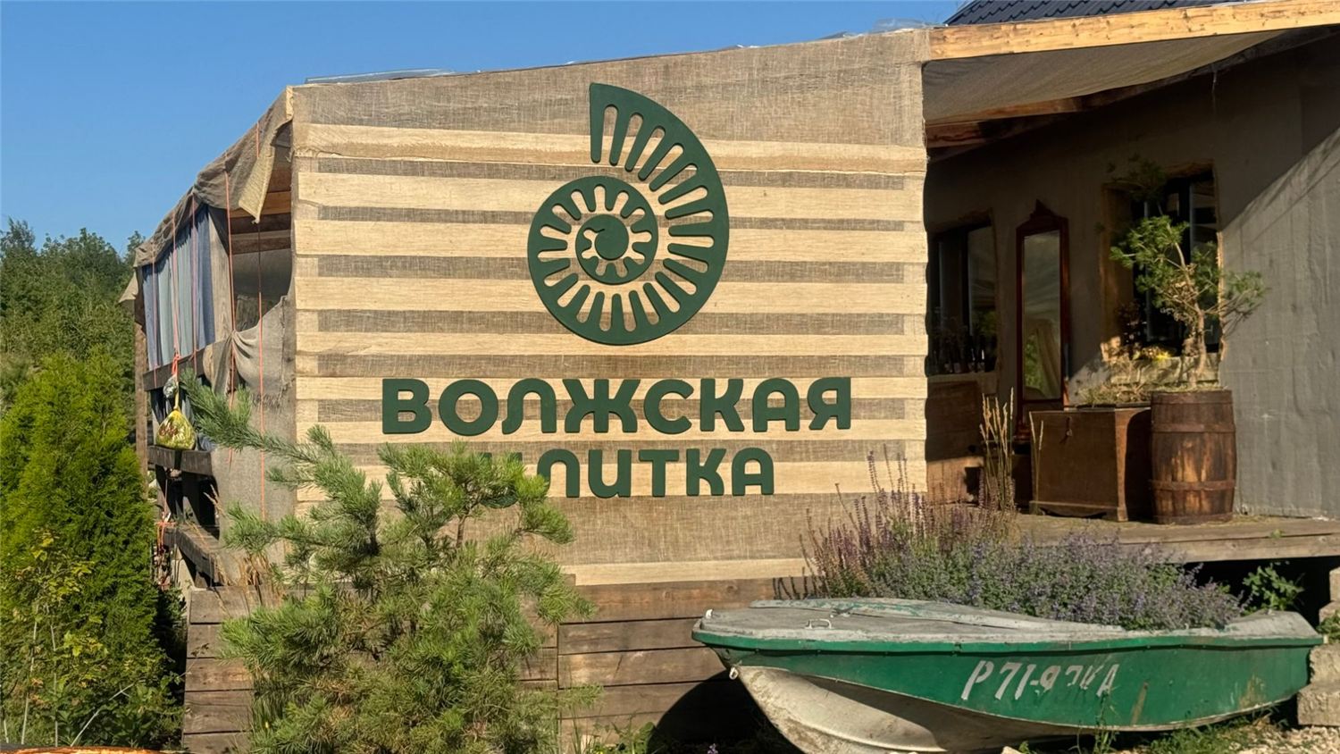 Вывеска «Волжская улитка» с ракушкой на стене фермы из мешковины, рядом старая лодка с цветами